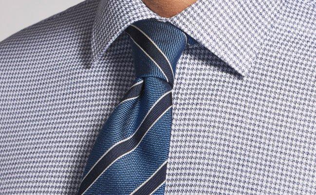 A close up of a model wearing a blue tie with black stripes, paired with a houndstooth casual shirt