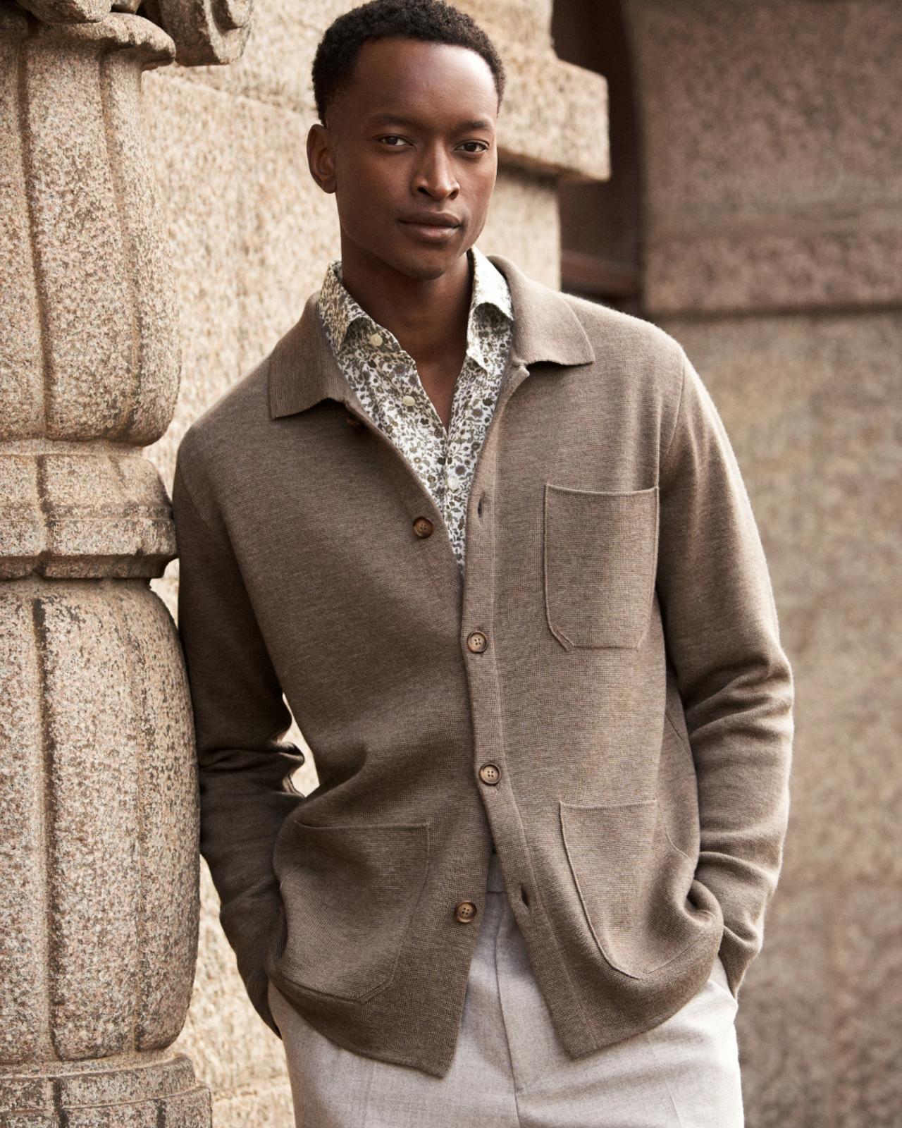 Man wearing a beige button-up cardigan over a patterned shirt and light trousers, standing against a stone wall