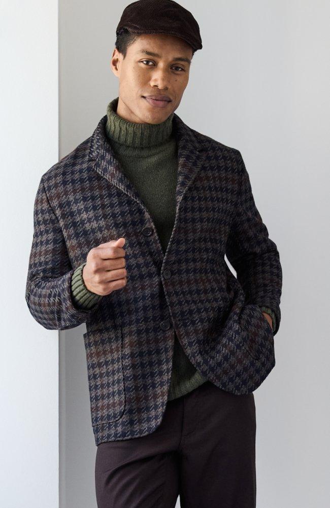 A male model wearing a dark grey check sport jacket paired with a dark green turtleneck sweater 