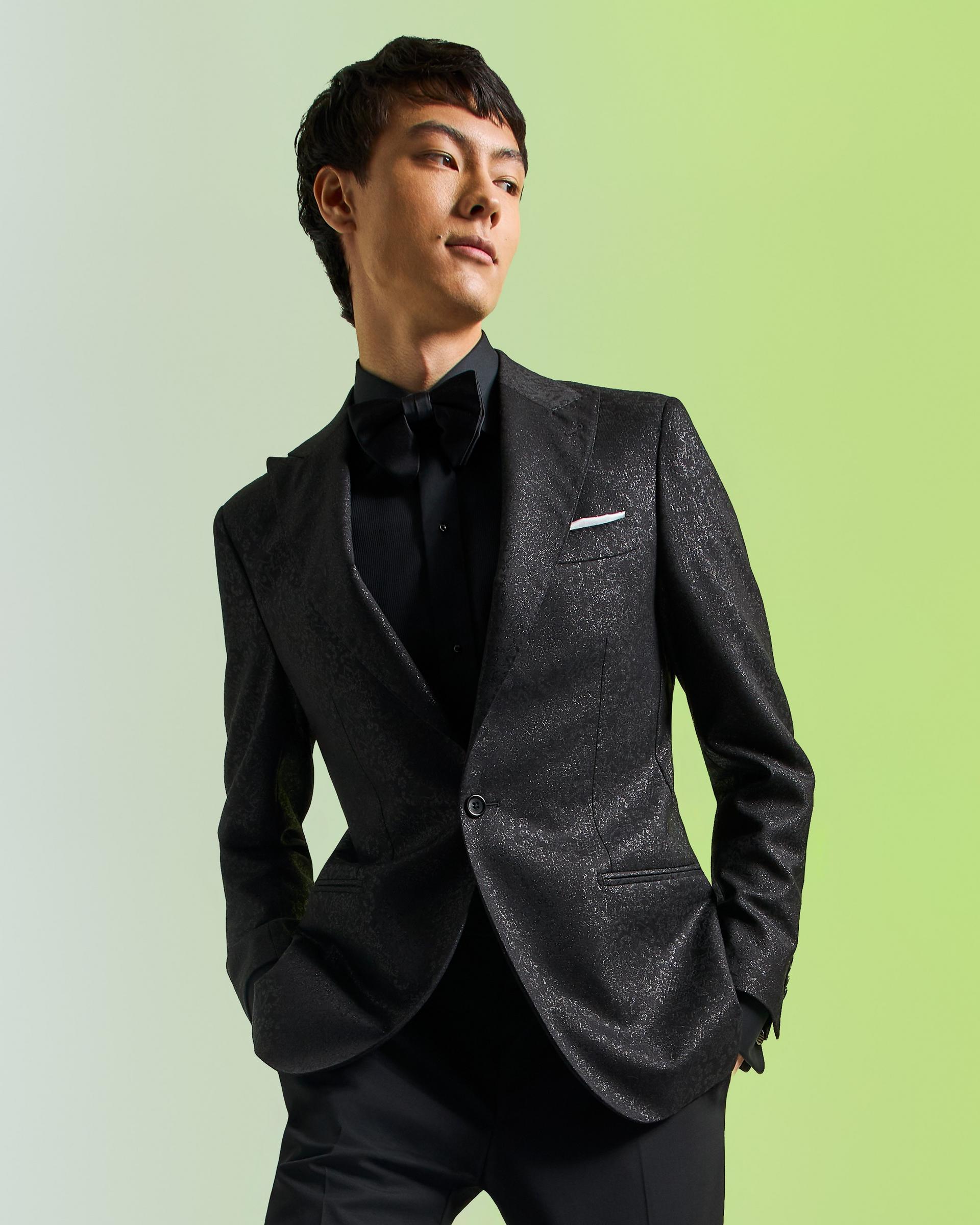 A male model wearing a black textured tailored Harold suit styled with a black bow tie and dress shirt