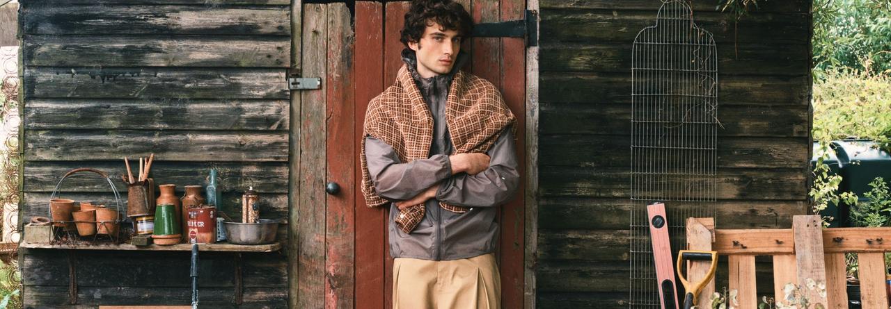 A model wearing beige pants and a grey jacket standing in front of a cottage