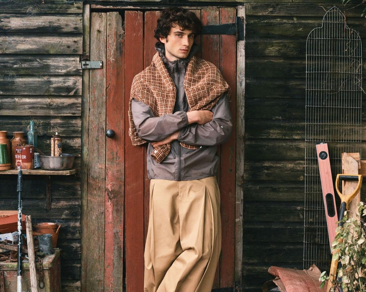 A model wearing beige pants and a grey jacket standing in front of a cottage