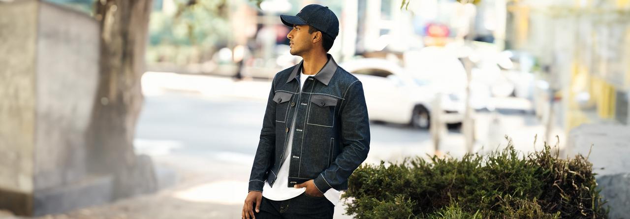 Homme portant une veste en denim foncé avec une casquette assortie, superposée sur un t-shirt blanc et un jean foncé, debout à l’extérieur