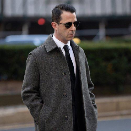 A man wearing a grey checkered overcoat over a suit and tie, paired with sunglasses.