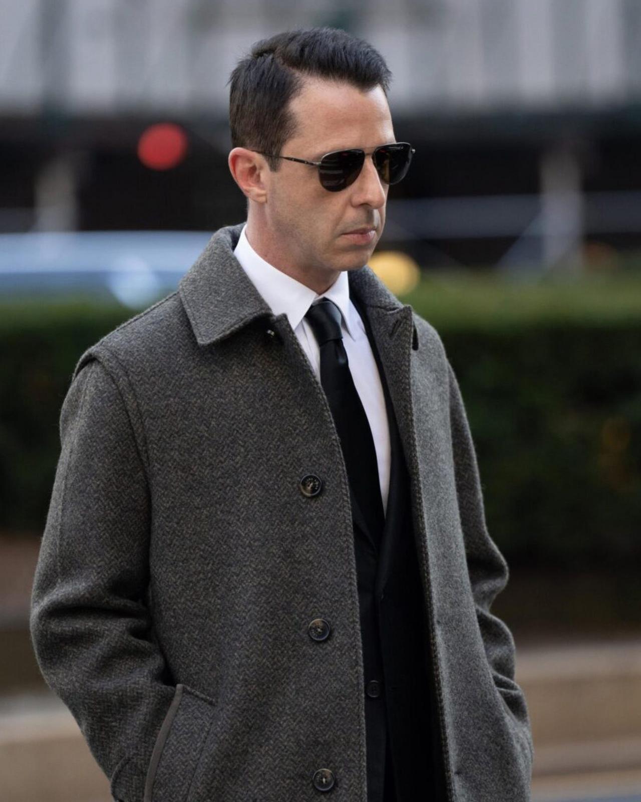 Kendall Roy de la série télé Succession, portant un manteau gris, une chemise blanche, une cravate noire et des lunettes de soleil