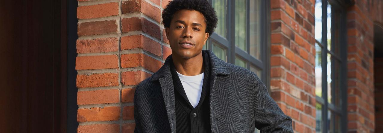 Un mannequin masculin porte un manteau gris, un cardigan noir et un t-shirt blanc, assortis d’un pantalon noir