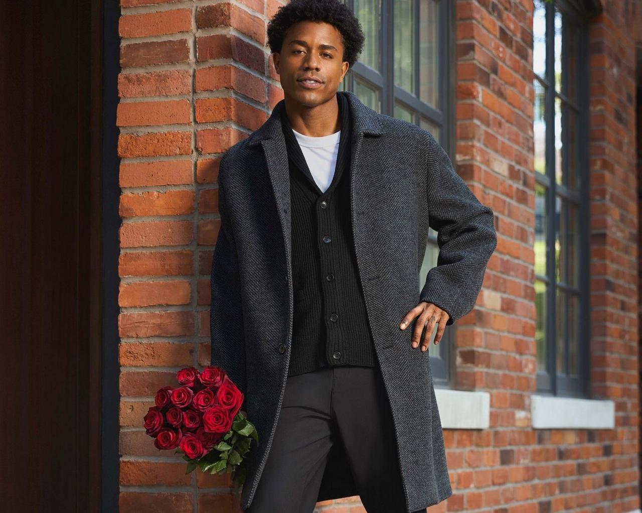 A male model wearing a grey coat, black cardigan, and white t-shirt paired with black trousers