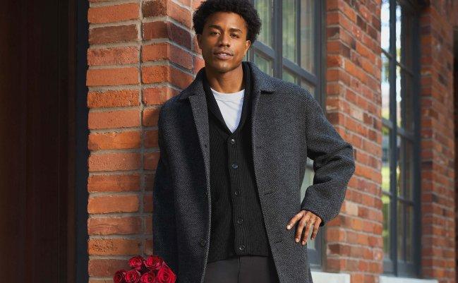 A male model wearing a grey coat, black cardigan, and white t-shirt paired with black trousers