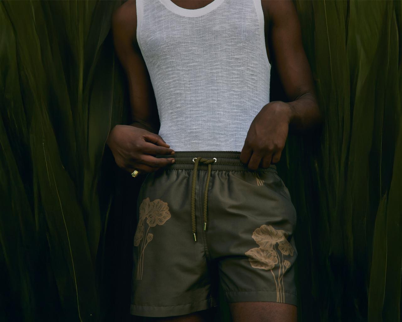 A model standing at a cottage  wearing white tank top and an olive floral printed shorts
