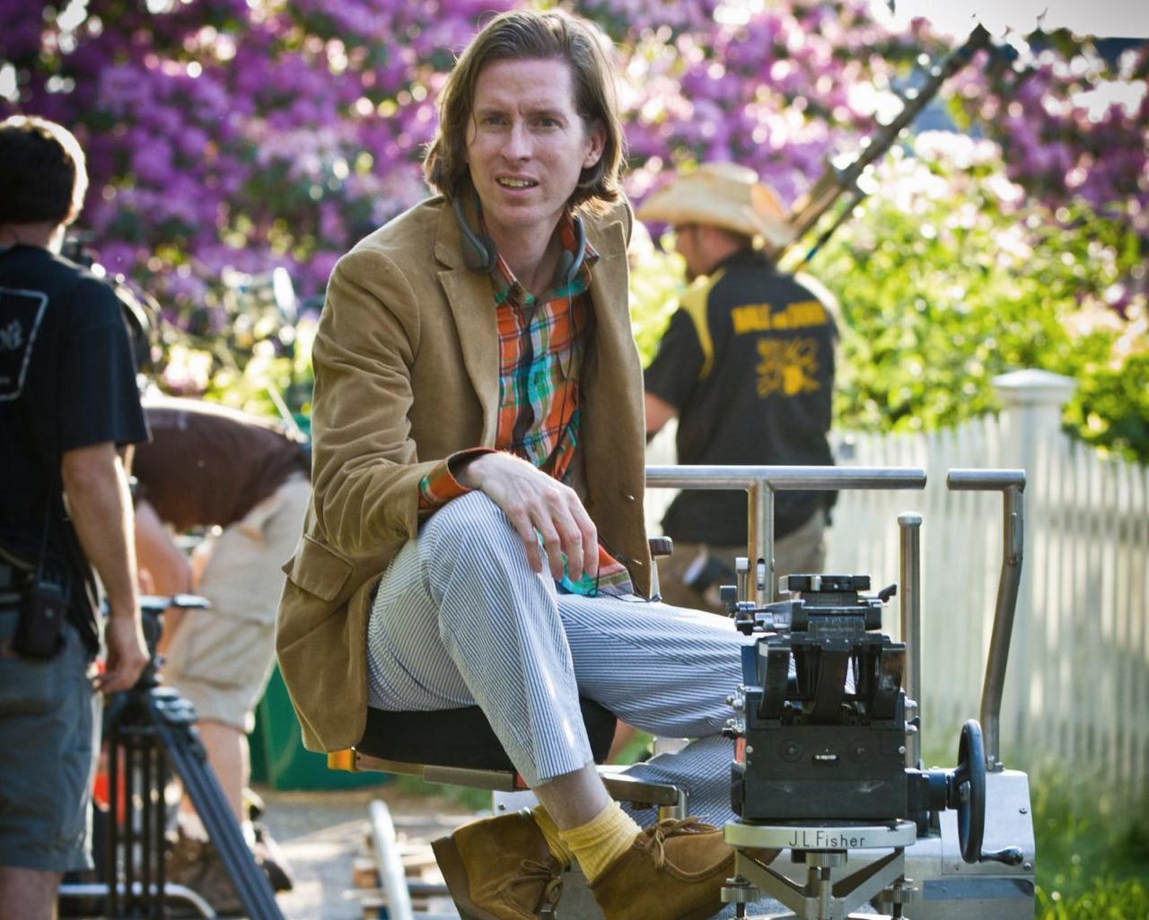 Wes Anderson avec des mocassins en suède beige, des chaussettes jaunes, un pantalon rayé, une chemise à carreaux et un veston brun
