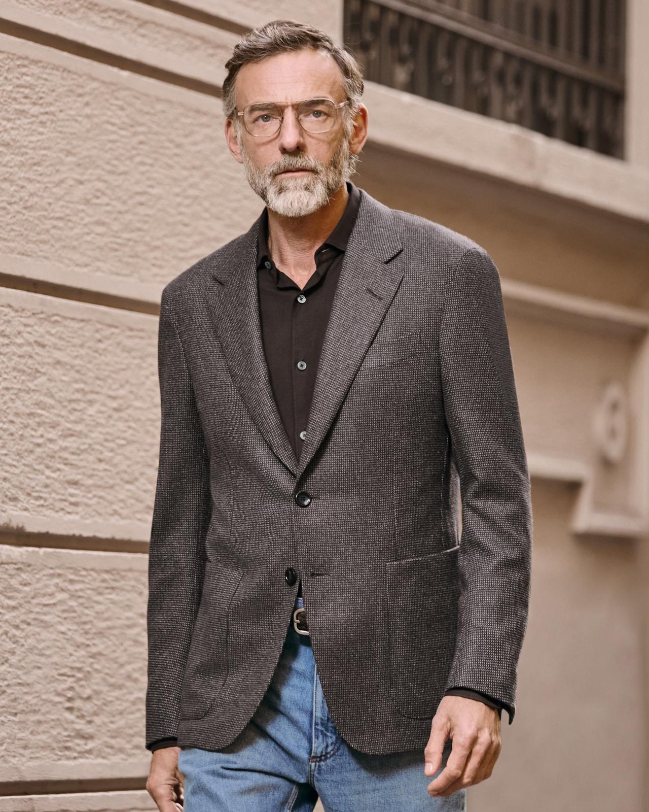 Man wearing a dark grey blazer over a black shirt paired with blue jeans, standing outdoors against a beige wall