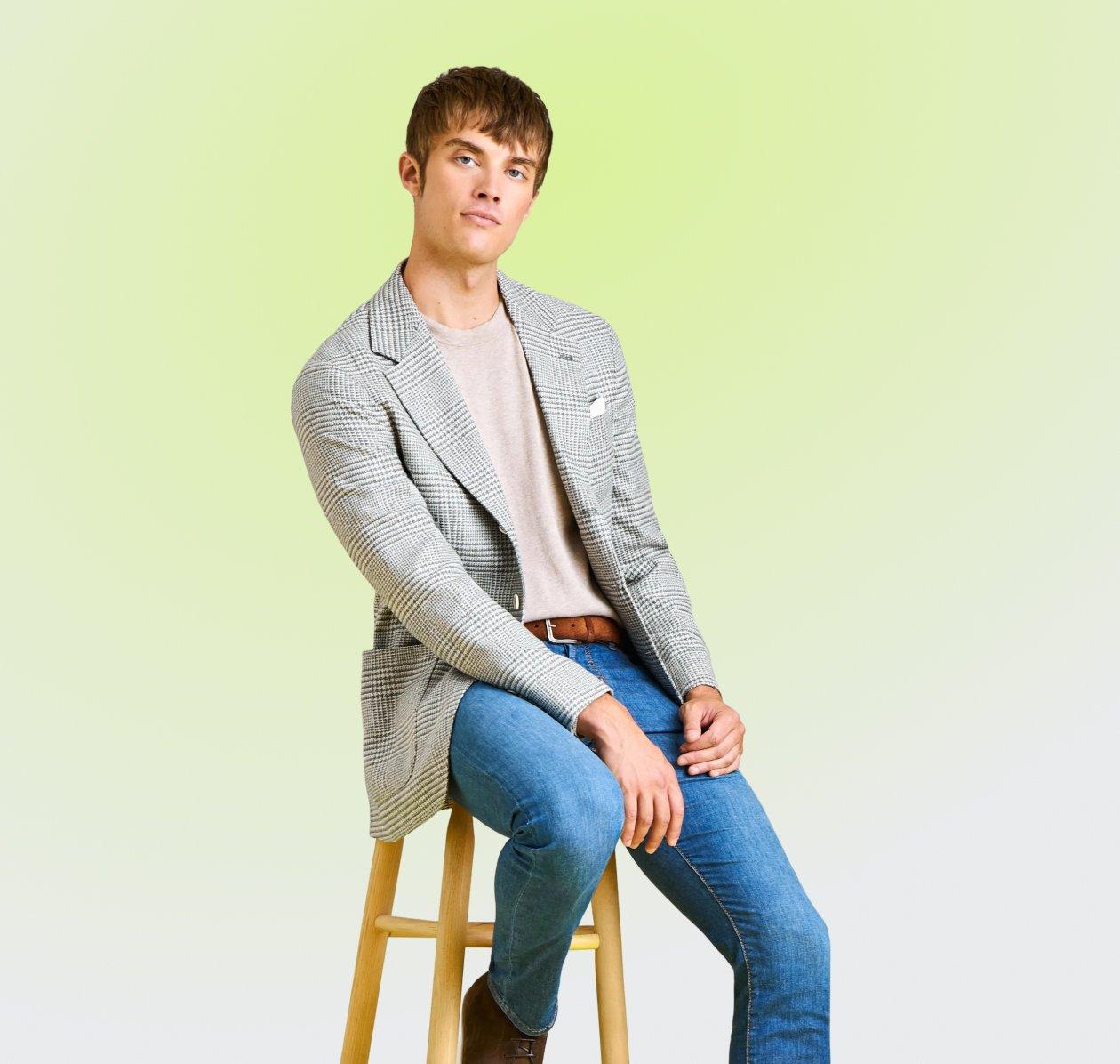 A man seated on a stool, wearing a light gray checkered blazer over a beige crew-neck shirt paired with blue jeans