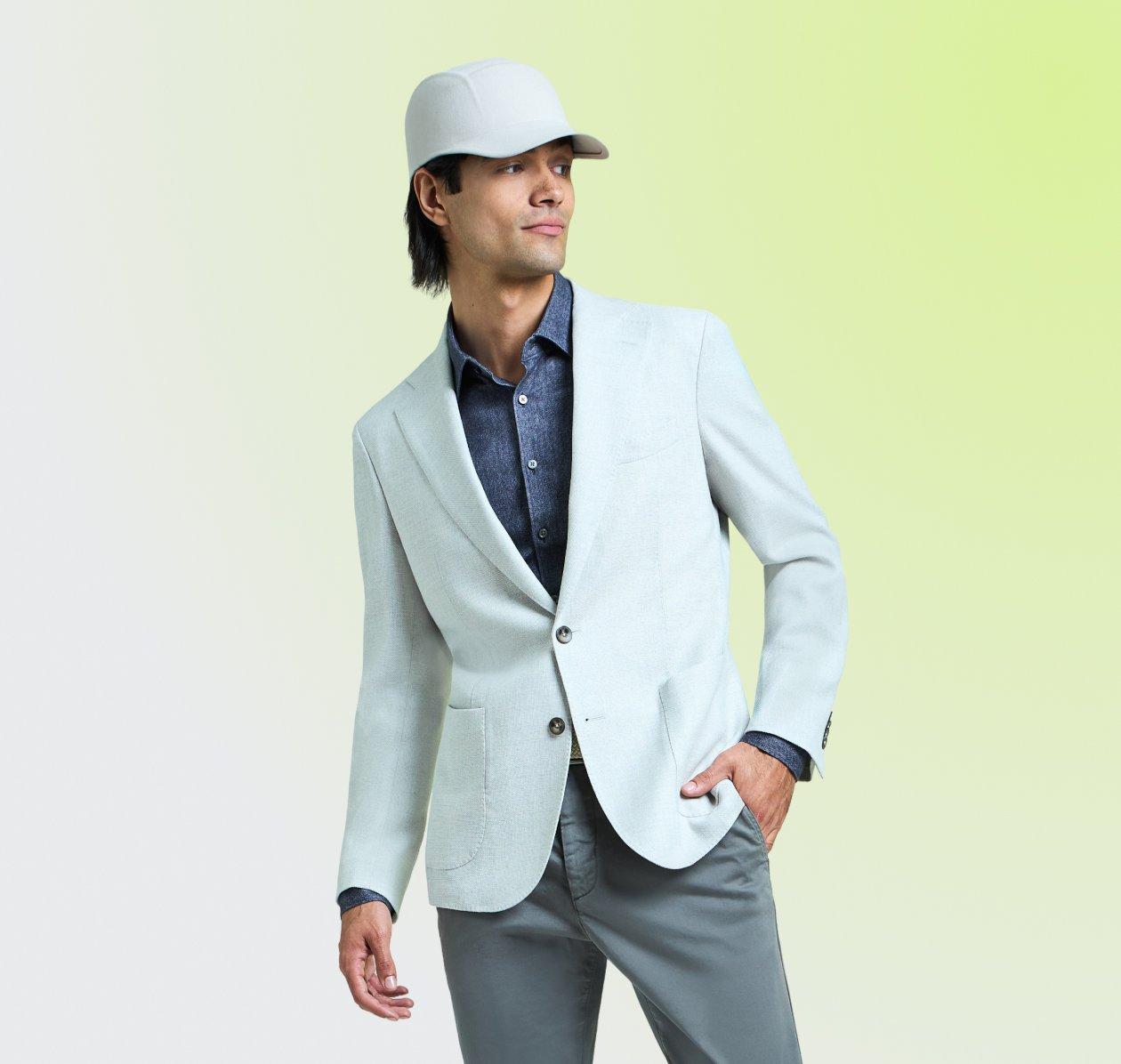 Homme portant un veston bleu pâle, une chemise boutonnée bleue, un pantalon gris et une casquette de baseball blanche.