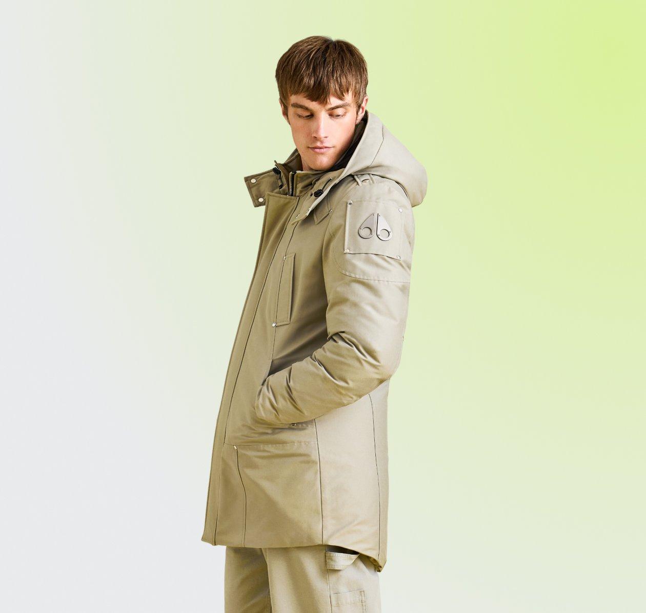 Man wearing a beige hooded parka with front pockets, standing against a light green background.