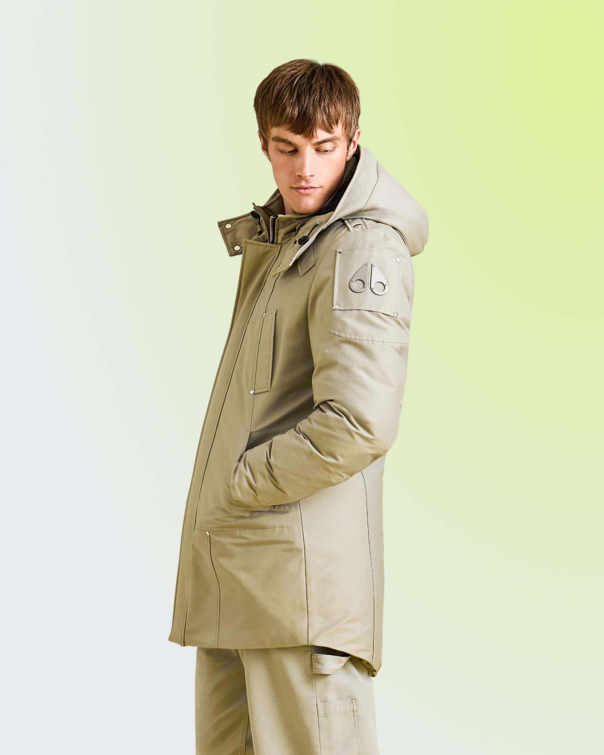 Man wearing a beige hooded parka with front pockets, standing against a light green background.