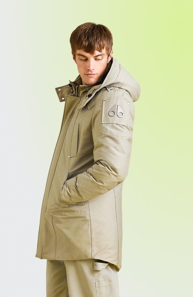 Homme portant une parka beige à capuchon avec des poches avant, debout sur un fond vert pâle.