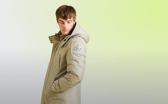 Man wearing a beige hooded parka with front pockets, standing against a light green background.