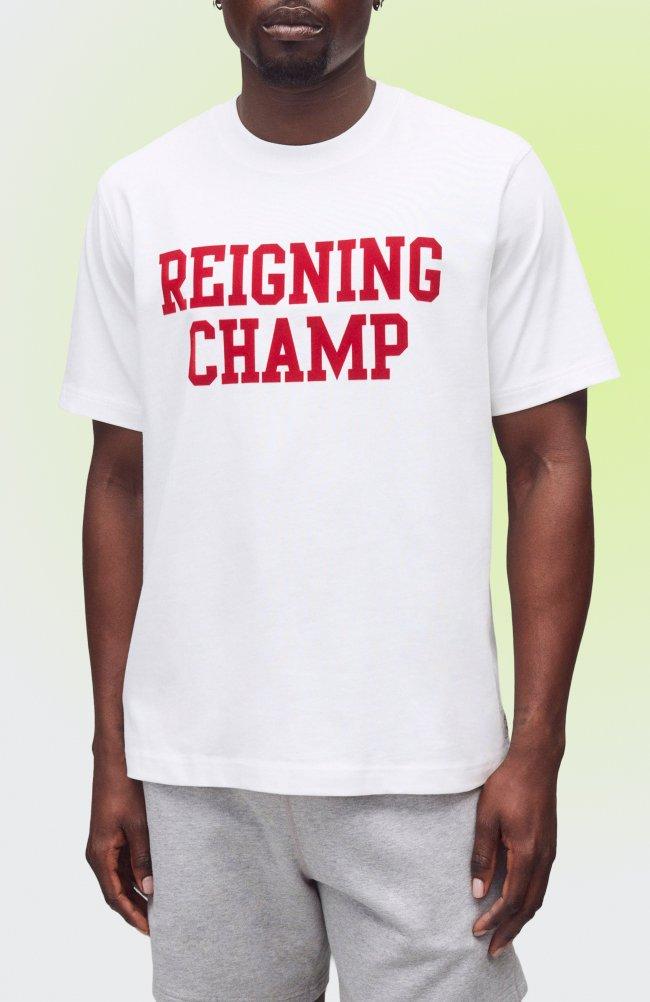 Un homme portant un T-shirt blanc avec l’inscription rouge « REIGNING CHAMP » et un pantalon de survêtement gris