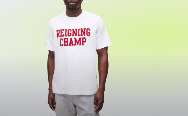 Man wearing a white T-shirt with red “REIGNING CHAMP” lettering and grey sweatpants