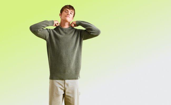Man wearing a green ribbed sweater and beige trousers, standing against a light green background.