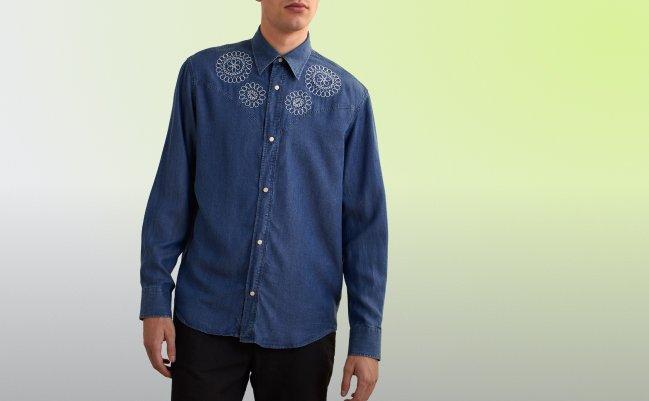Un homme portant une chemise boutonnée en denim bleu, ornée d’une broderie blanche circulaire complexe sur la poitrine