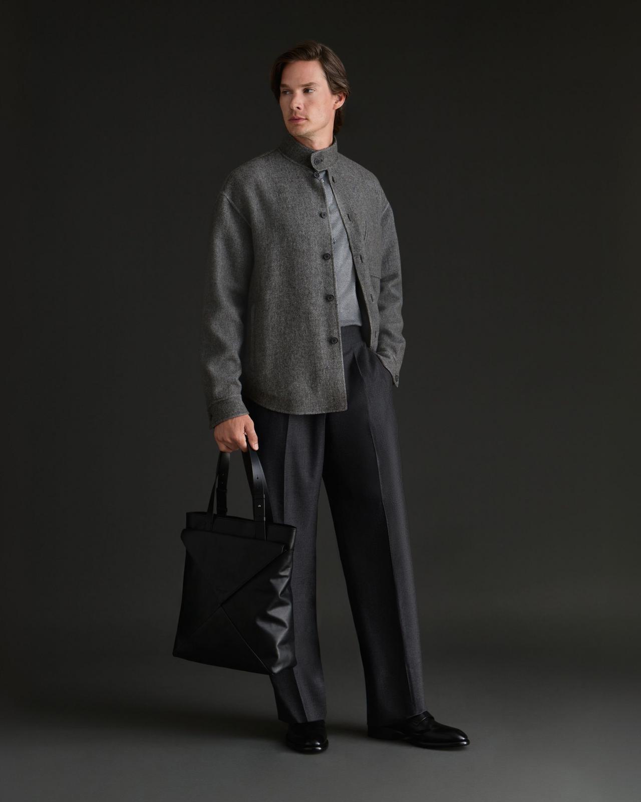 Un homme portant une veste grise cintrée sur une chemise gris pâle, agencée avec un pantalon foncé, et tenant un élégant sac fourre-tout noir