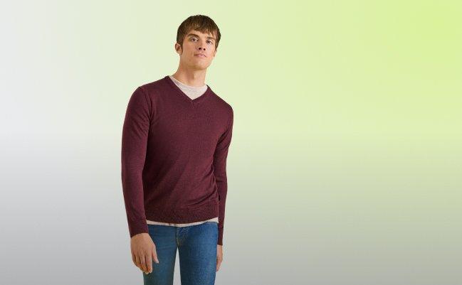 Man wearing a burgundy V-neck sweater over a beige T-shirt and blue jeans