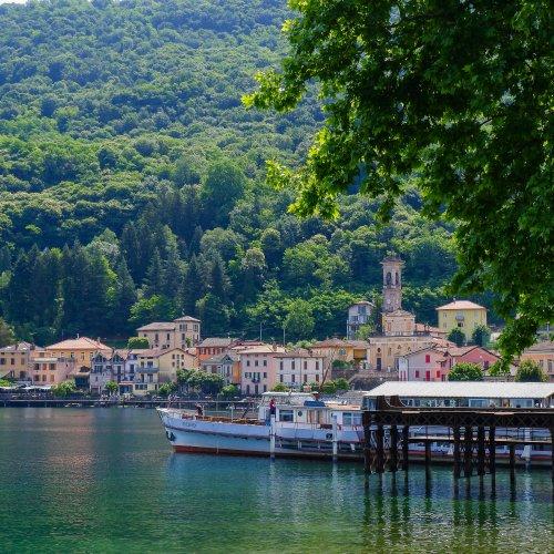 Village italien au bord d'un lac avec des bâtiments colorés et un clocher, bateaux amarrés à un quai, Made in Italy, A/H26