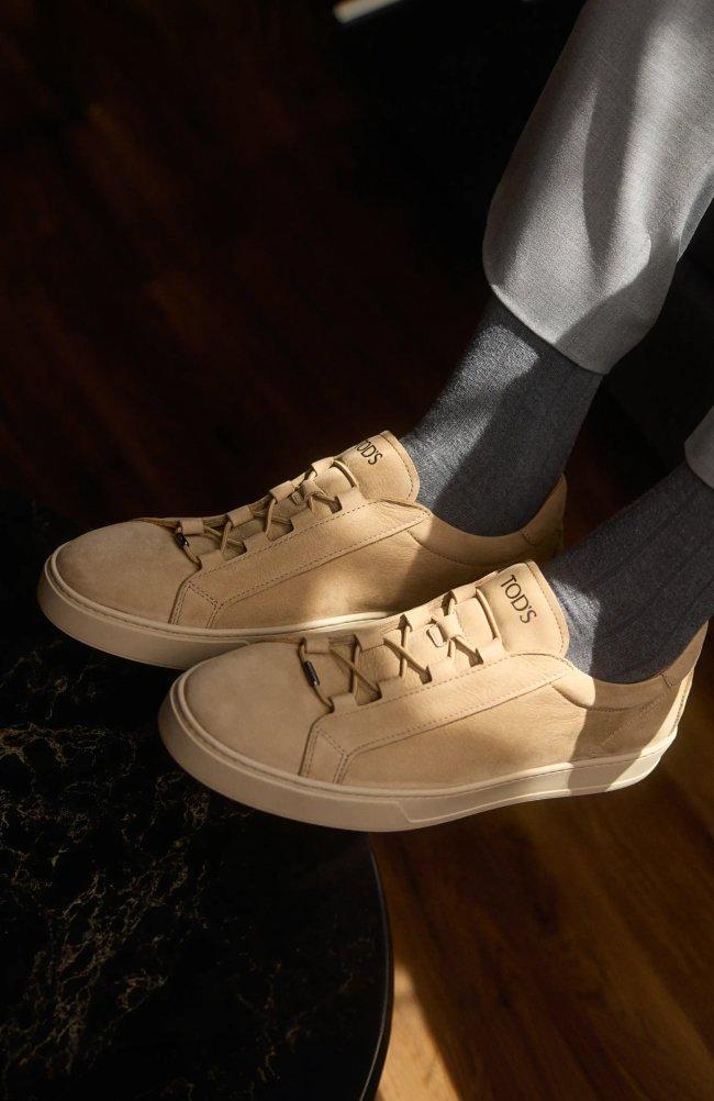 Beige leather sneakers with laces worn with grey socks and light grey trousers.