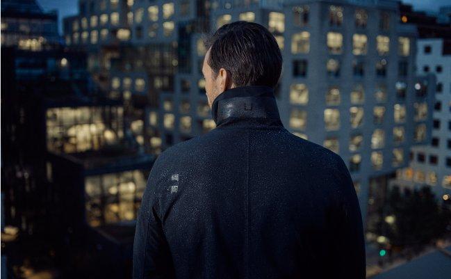 Homme vu de dos portant une veste UBR noire, surplombant les gratte-ciel d'une ville la nuit, collection A/H26