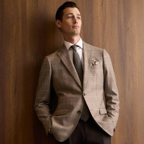 Un homme portant un blazer beige à carreaux bien ajusté, accompagné d’une chemise habillée blanche, d’une cravate foncée et d’un mouchoir de poche décoratif