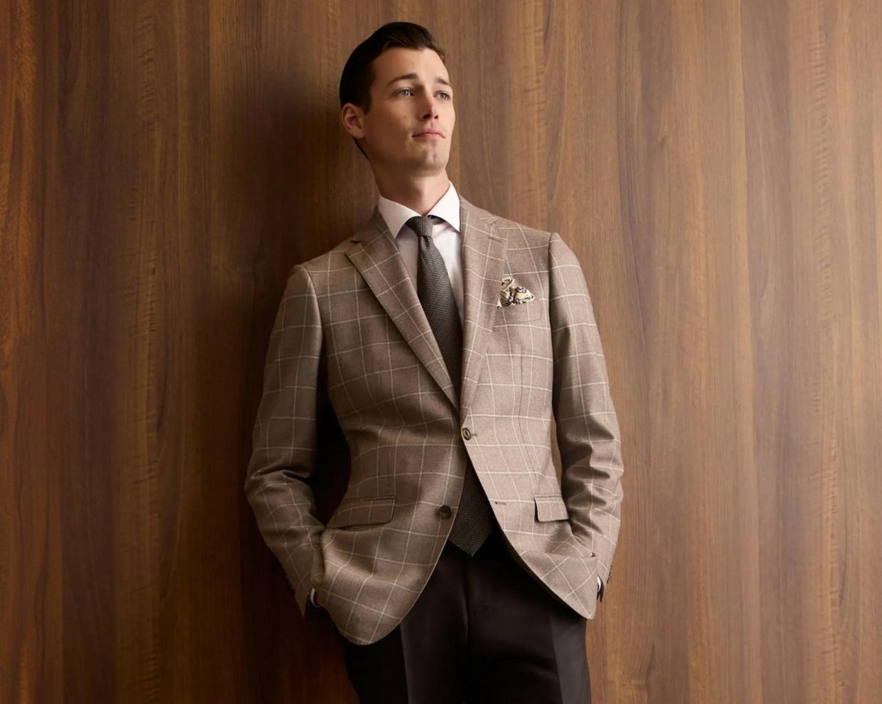 Un homme portant un blazer beige à carreaux bien ajusté, accompagné d’une chemise habillée blanche, d’une cravate foncée et d’un mouchoir de poche décoratif