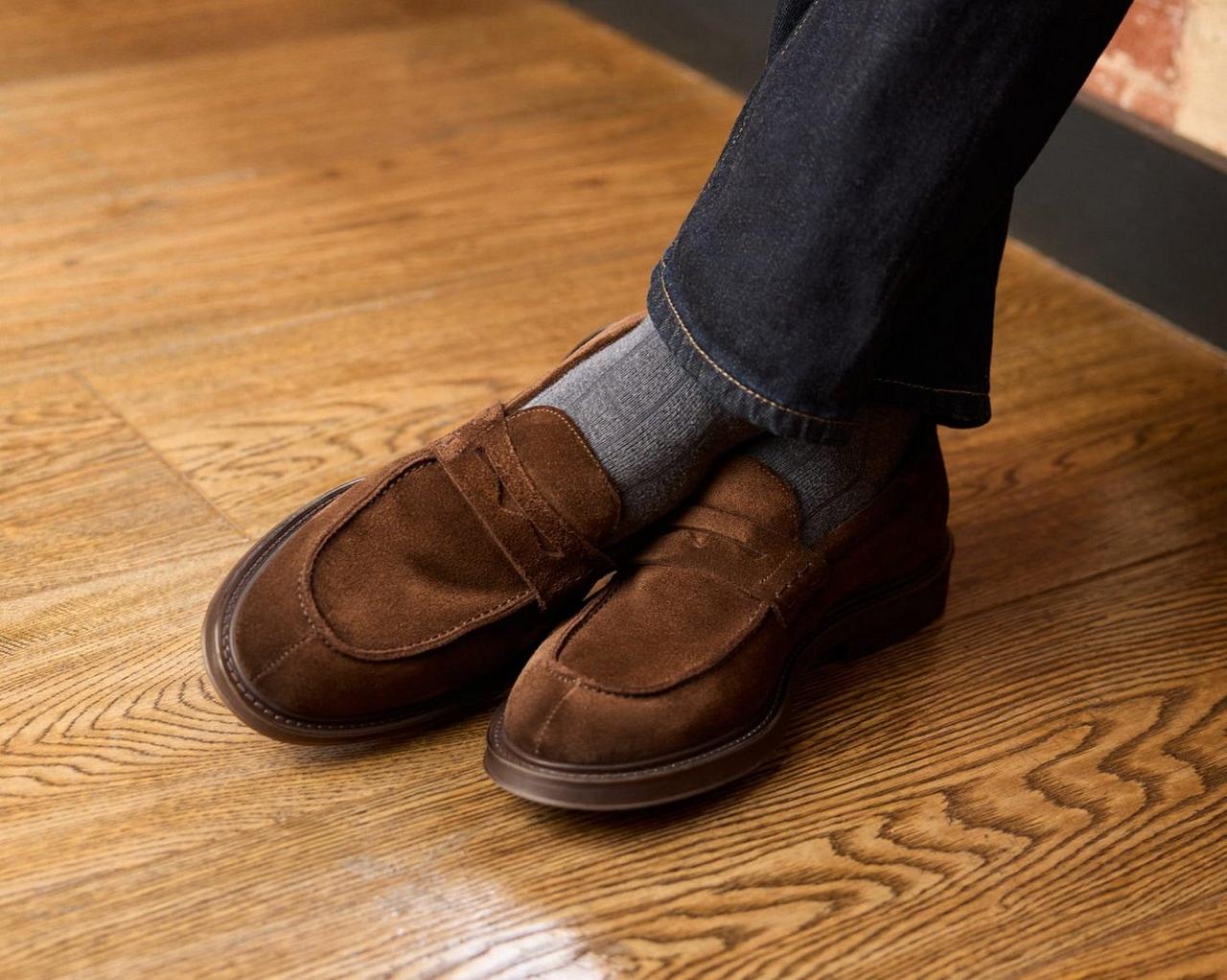 Une paire de mocassins en suède brun portés avec des chaussettes grises et un jean foncé, debout sur un plancher en bois poli