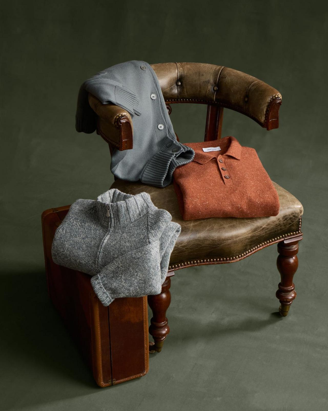 Three folded knit pieces, a grey button cardigan, a rust polo sweater, and a grey marled quarter-zip, styled on a vintage leather chair, Knitwear, FW26 collection