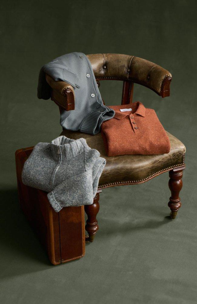 Three folded knit pieces, a grey button cardigan, a rust polo sweater, and a grey marled quarter-zip, styled on a vintage leather chair, Knitwear, FW26 collection