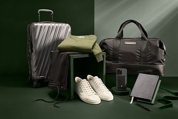 Baskets blanches Prada, coque d'iPhone, carnet gris et stylo, pull vert, valise Tumi et pantalons noirs