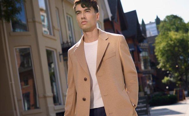 A male model wearing a tan colored BOSS wool-cashmere coat, white T-shirt, and black jeans paired with a brown leather briefcase