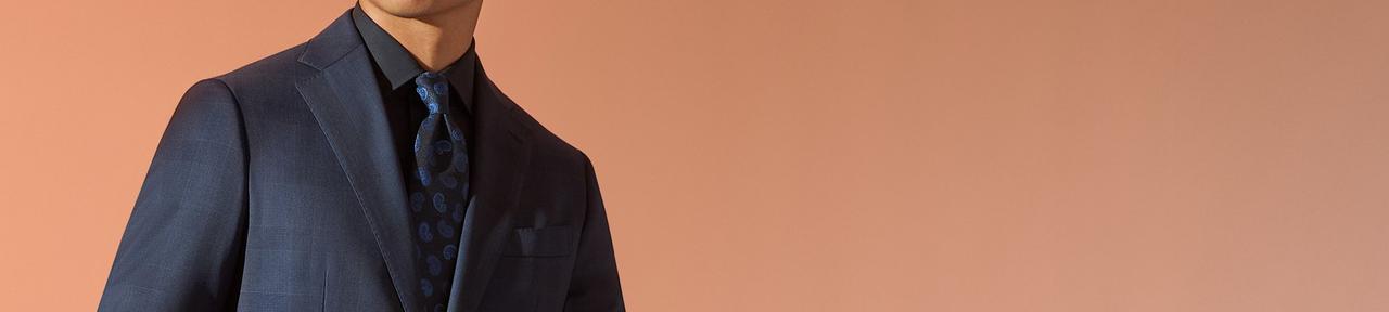 A close up of a male model wearing a navy fine tailored suit paired with a black and navy paisley tie