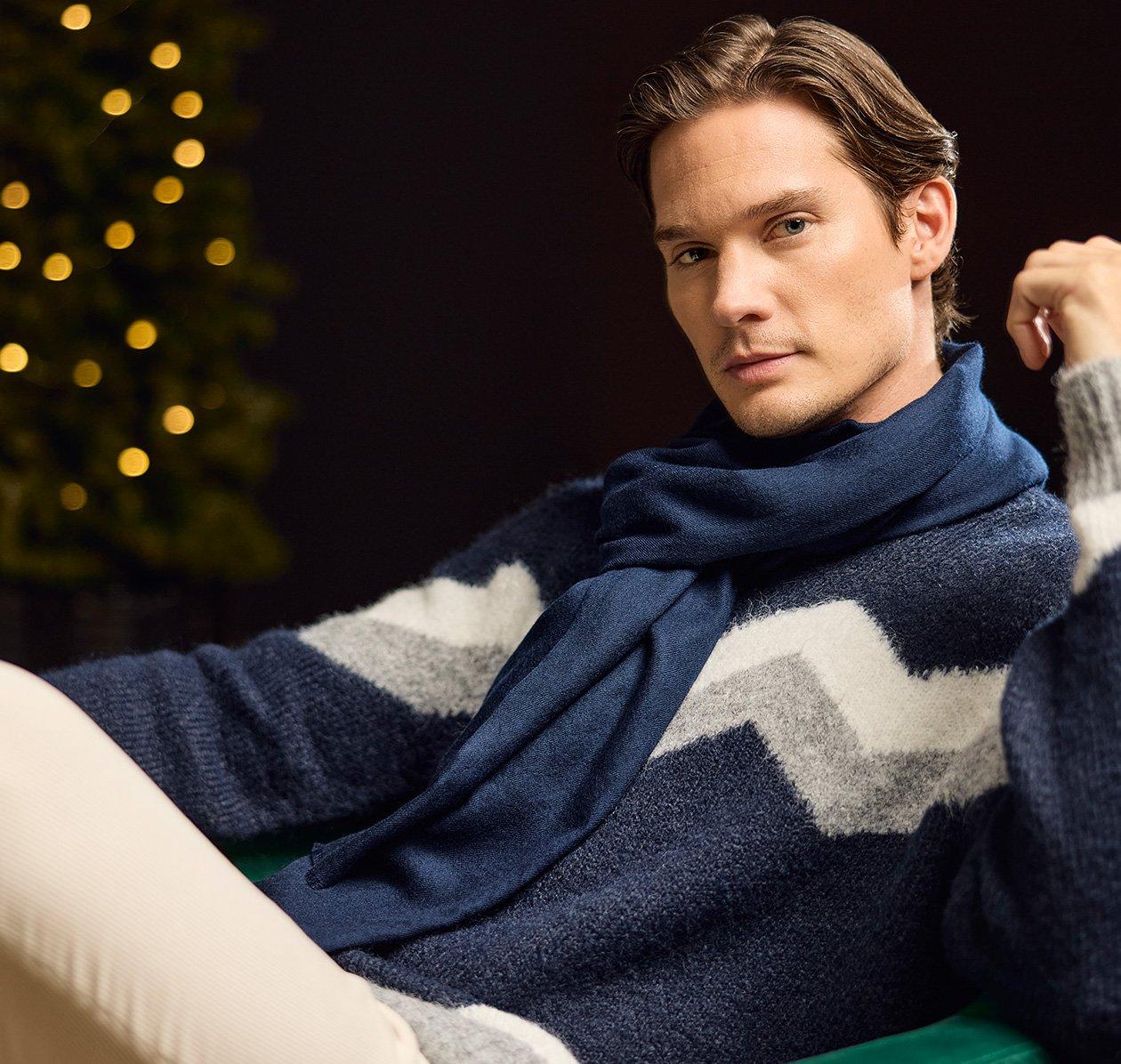 A male model wearing a blue knit sweater with a grey and white chevron pattern, paired with a blue knit scarf