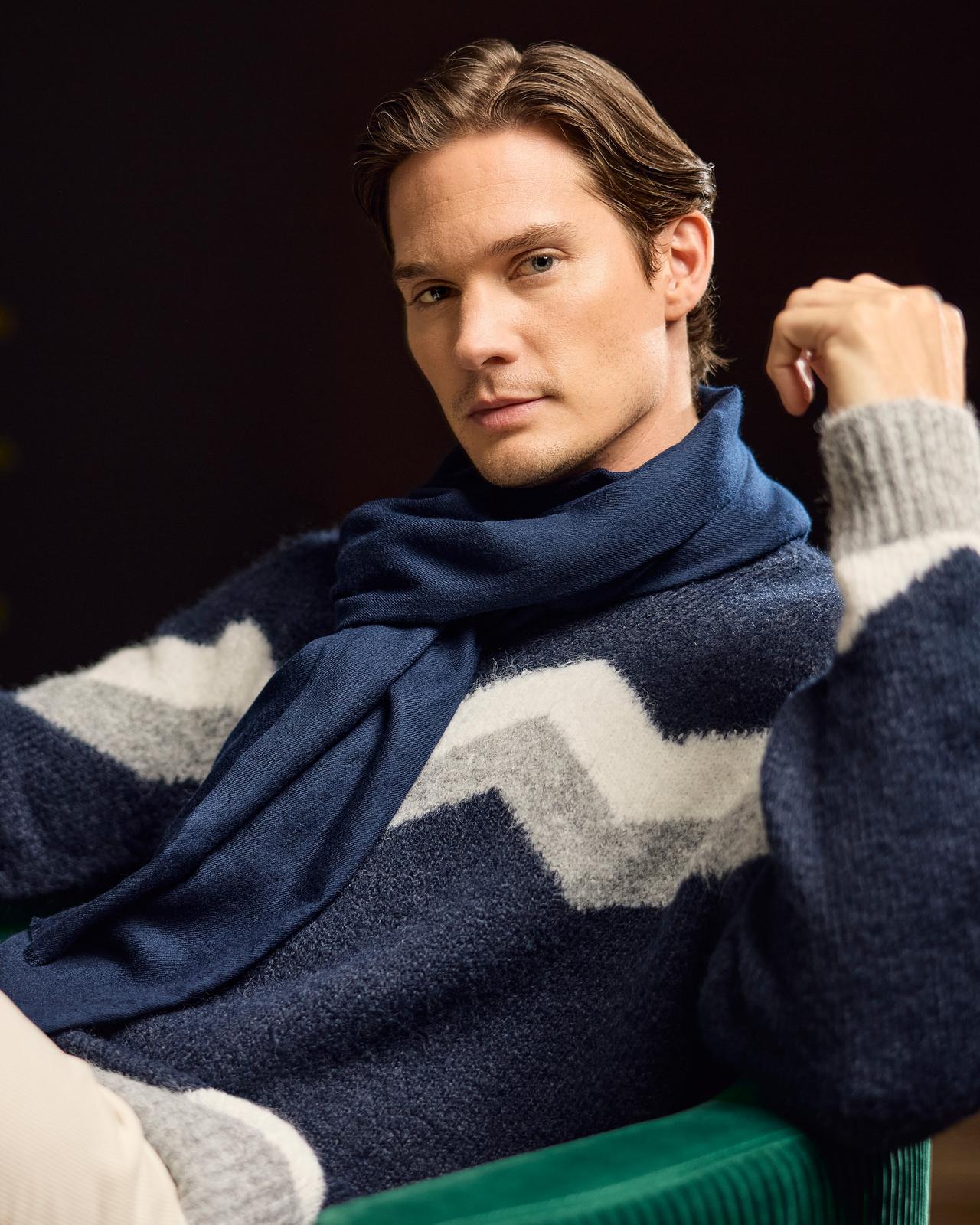 Un mannequin homme portant un pull en tricot bleu avec un motif chevron gris et blanc, assorti d’une écharpe en tricot bleue