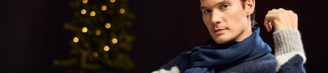 A male model wearing a blue knit sweater with a grey and white chevron pattern, paired with a blue knit scarf