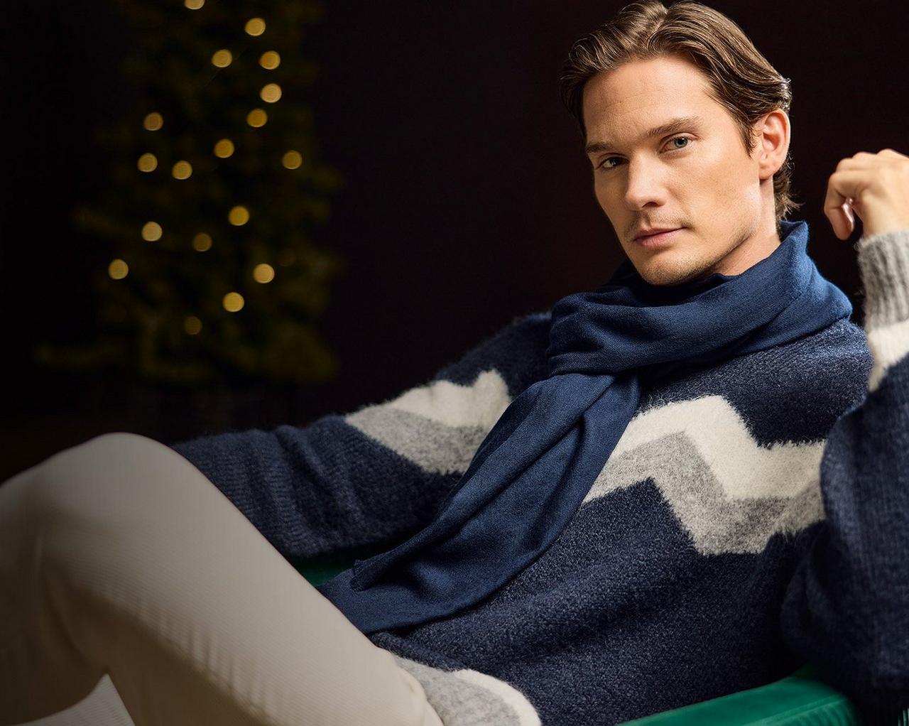 Un mannequin homme portant un pull en tricot bleu avec un motif chevron gris et blanc, assorti d’une écharpe en tricot bleue
