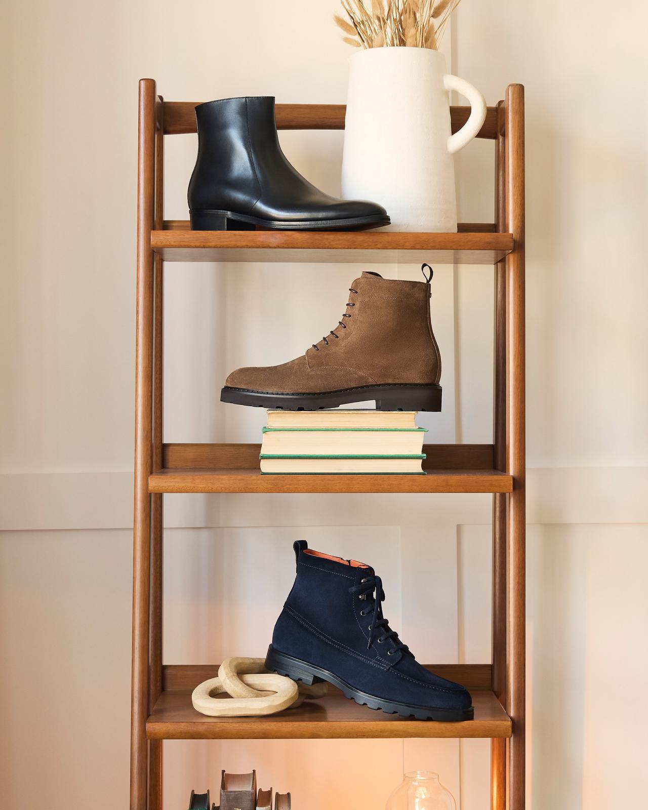 Three boot styles on a shelf; a black leather ankle boot, a brown suede ankle lug boot, and a black suede lace-up boot