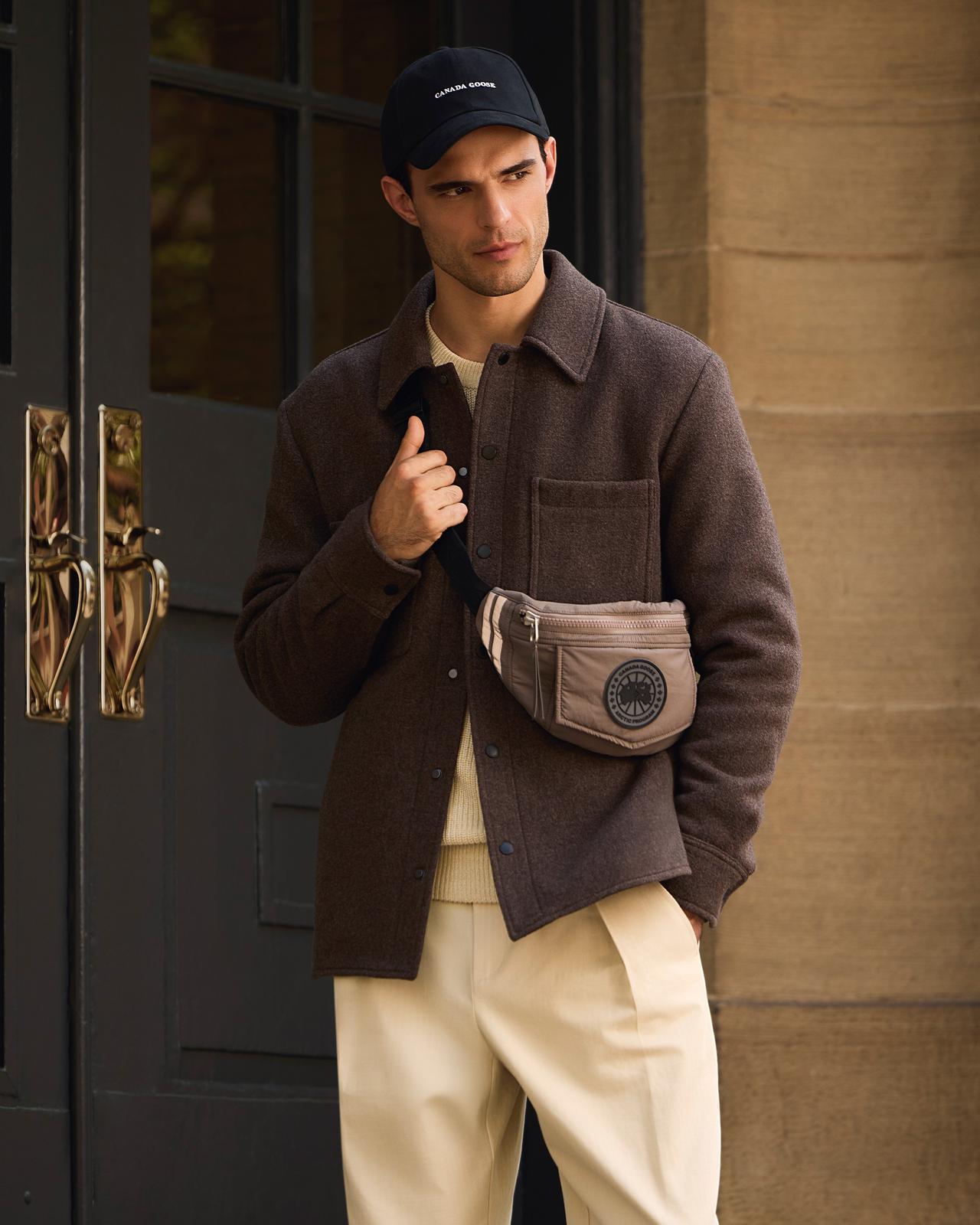 A male model wearing a brown Canada Goose shirt jacket, paired with tan colored chino pants, a light brown waist pack and black cap