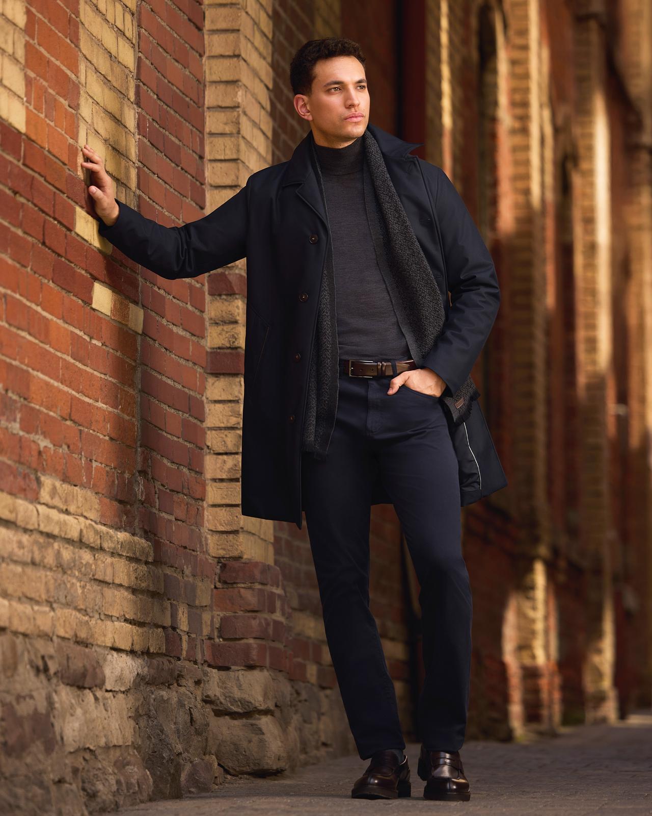 A male model wearing a navy Canali lightweight jacket, dark grey mock neck wool sweater, and navy pants, paired with a brown leather belt and leather boots