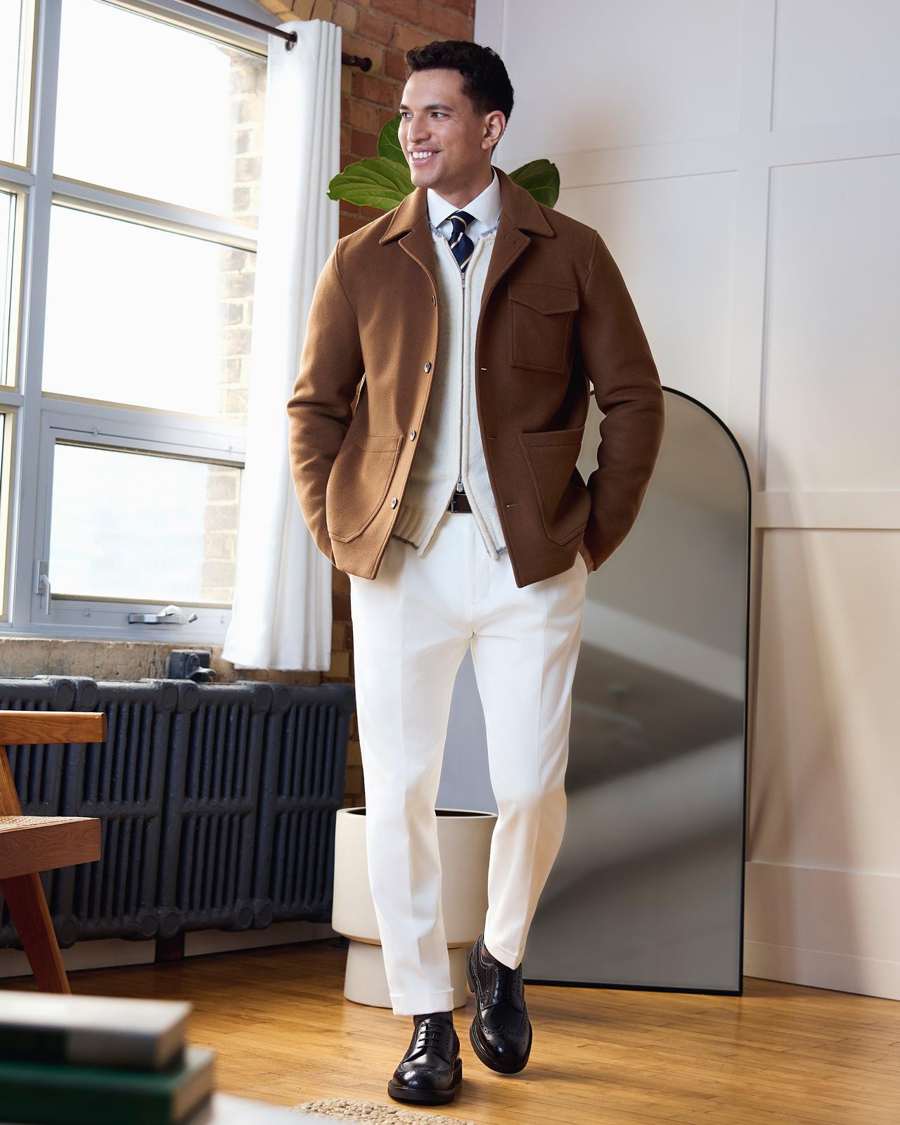A male model wearing a brown Herno jacket, paired with a cream colored zip-up knit sweater, white casual shirt, striped navy tie, white trousers, and black leather shoes