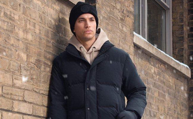 A male model wearing a grey suede heavyweight jacket with shearling collar paired with a beige knit zip up sweater