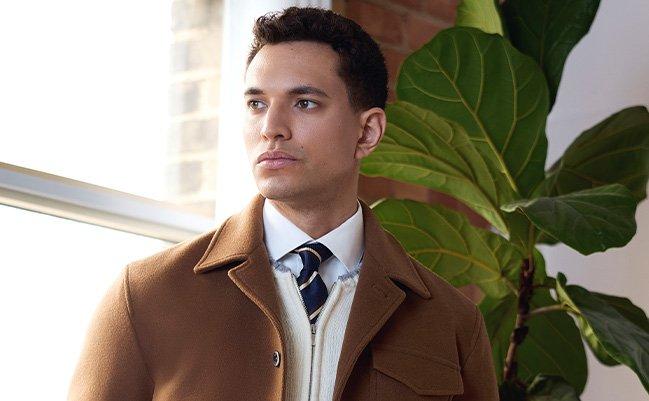 A male model wearing a brown lightweight jacket, paired with a cream colored zip-up knit sweater, white casual shirt, and striped navy tie