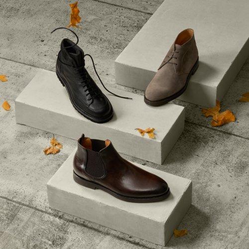 Three pairs of men’s leather shoes in black, brown, and dark brown displayed on concrete pedestals with scattered autumn leaves.