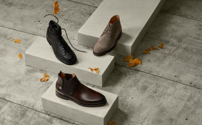 Trois paires de chaussures en cuir pour hommes, noires, brunes et brun foncé, présentées sur des socles en béton avec des feuilles d’automne éparses